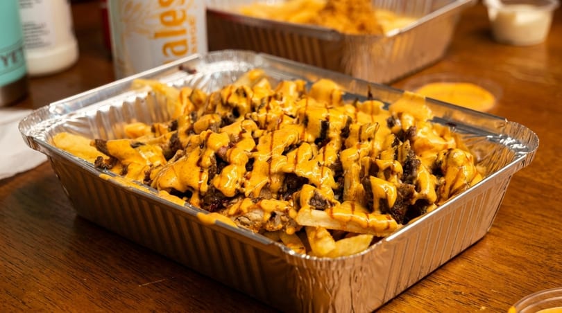 Fries topped with meat and cheese sauce in a foil container on a wooden table.