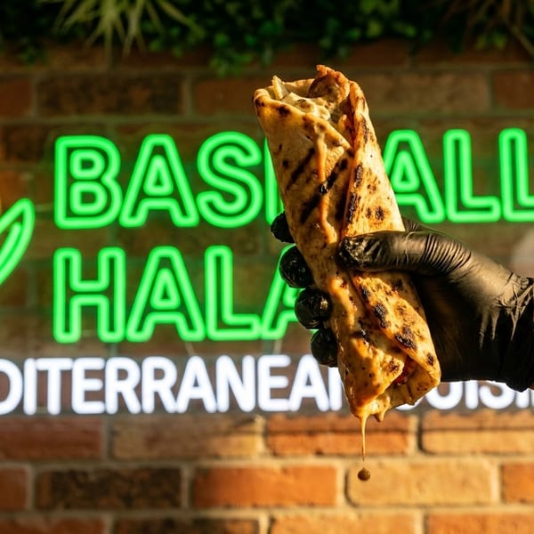 Hand in black glove holding a grilled wrap with sauce dripping, neon sign in background.