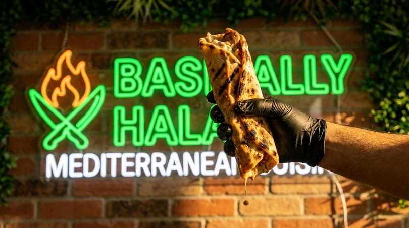 Hand in black glove holding a grilled wrap with sauce dripping, neon sign in background.