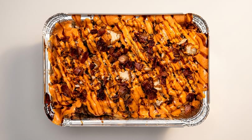 Loaded fries with chicken, bacon, and orange sauce in a foil container.
