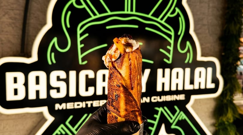 A hand in a black glove holds a dessert wrap topped with cream and fruit in front of a neon sign.