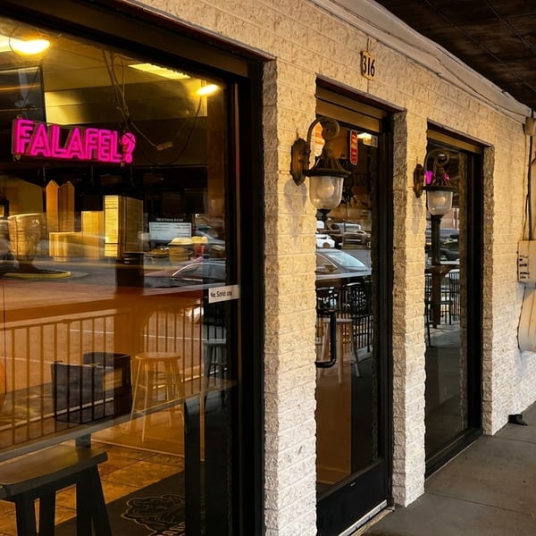 Exterior of a restaurant with a neon pink "FALAFEL?" sign visible through the window at dusk.
